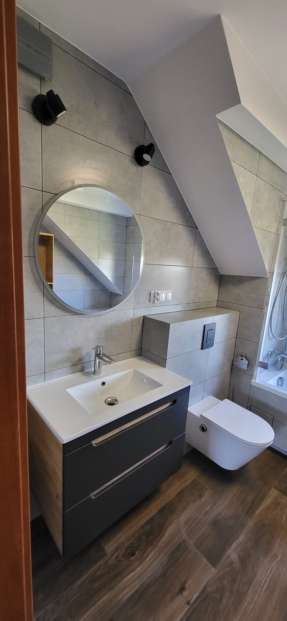 Nowoczesna łazienka na poddaszu z szarymi płytkami, okrągłym lustrem i drewnopodobną podłogą. Uwagę zwraca podwieszana toaleta i szafka z umywalką.