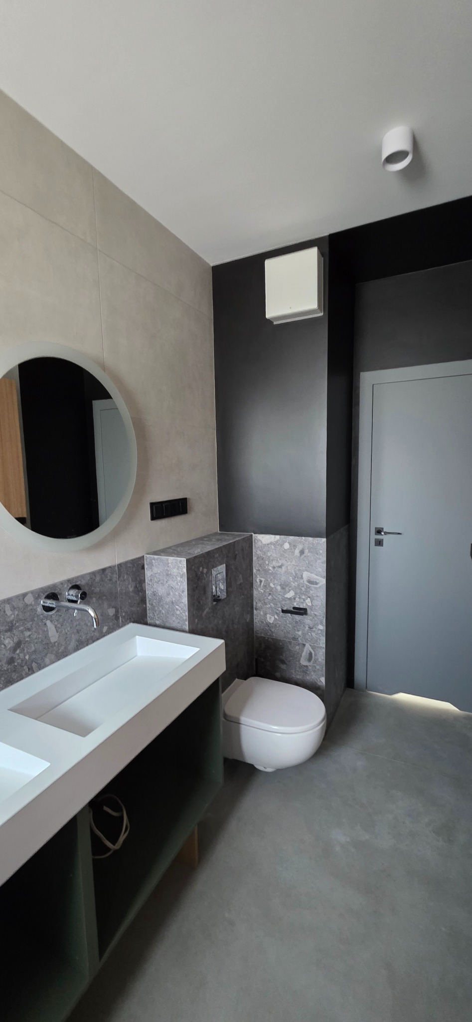 Nowoczesna łazienka z umywalką, okrągłym lustrem i toaletą w zabudowie. Szare płytki, czarna ściana i szare drzwi tworzą spójną kolorystykę.
