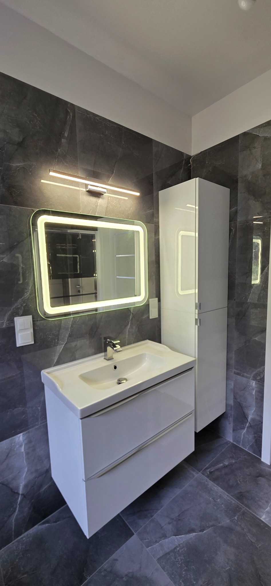 Nowoczesna łazienka z szarymi płytkami imitującymi marmur, białą szafką z umywalką i podświetlanym lustrem. Minimalistyczny design, eleganckie wykończenie.