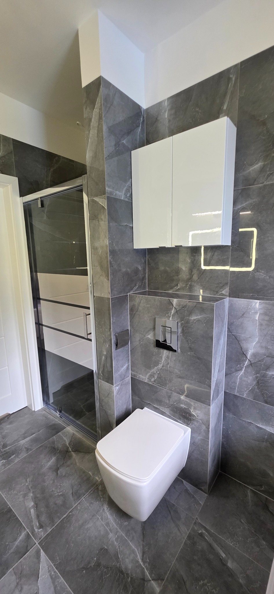 Elegancka, nowoczesna łazienka z szarą marmurową okładziną, podwieszaną toaletą i białą szafką. Widoczna kabina prysznicowa z elementami chromowanymi.
