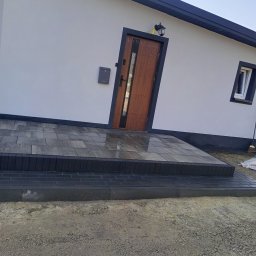 DAX-BUD Usługi remonowo -budowlane Daniel Worożbit - Ułożona kostka brukowa przed wejściem do domu z szarą elewacją i ciemną stolarką. Widoczny fragment elewacji z oknem i drzwiami wejściowymi.