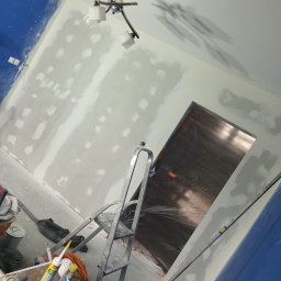 DAX-BUD Usługi remonowo -budowlane Daniel Worożbit - Wnętrze w trakcie szpachlowania ścian, widoczna drabina, zakryte folią drzwi, puszki z farbą i narzędzia. Ściany częściowo pomalowane na niebiesko.