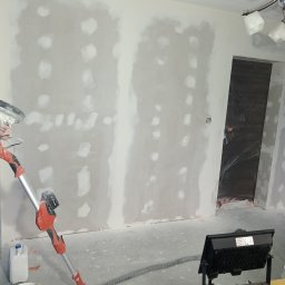 DAX-BUD Usługi remonowo -budowlane Daniel Worożbit - Szlifowanie ściany szlifierką z odkurzaczem, widoczne załatane dziury i nierówności. W tle zabezpieczone folią drzwi i lampa budowlana. Etap przygotowania do malowania.