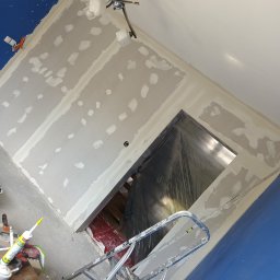 DAX-BUD Usługi remonowo -budowlane Daniel Worożbit - Wnętrze w trakcie szpachlowania ścian gipsowych, widoczna drabina, narzędzia i zabezpieczone folią drzwi. Ściany pomalowane na niebiesko, sufit biały, lampa sufitowa.