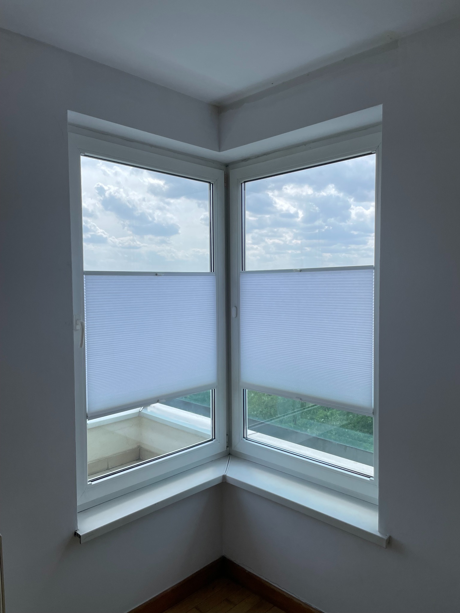 Białe plisy okienne zamontowane w narożnym oknie, widok na zachmurzone niebo z mieszkania w Poznaniu. Delikatne światło dzienne.