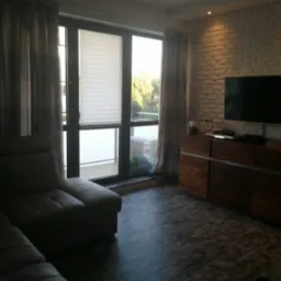 Stylowe wnętrze salonu z ceglaną ścianą, drewnianą komodą z telewizorem i wygodną sofą. Widok na balkon przez duże okno z roletami.