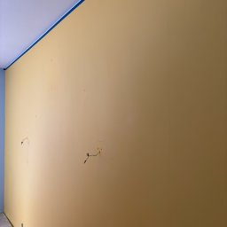 ABInterior - Żółta ściana po malowaniu, zabezpieczona taśmą malarską przy suficie i folią na podłodze. Widoczne gniazdka elektryczne i kable. Świeżo pomalowane wnętrze.