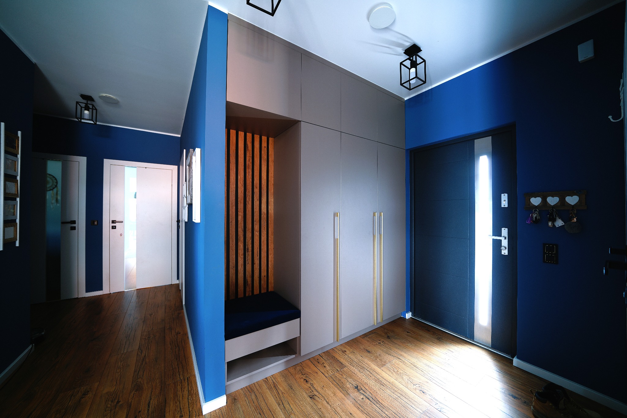 Nowoczesny przedpokój w odcieniach granatu i szarości z drewnianą podłogą. Wbudowana szafa z siedziskiem i pionowymi lamelami. Dwoje drzwi w tle.