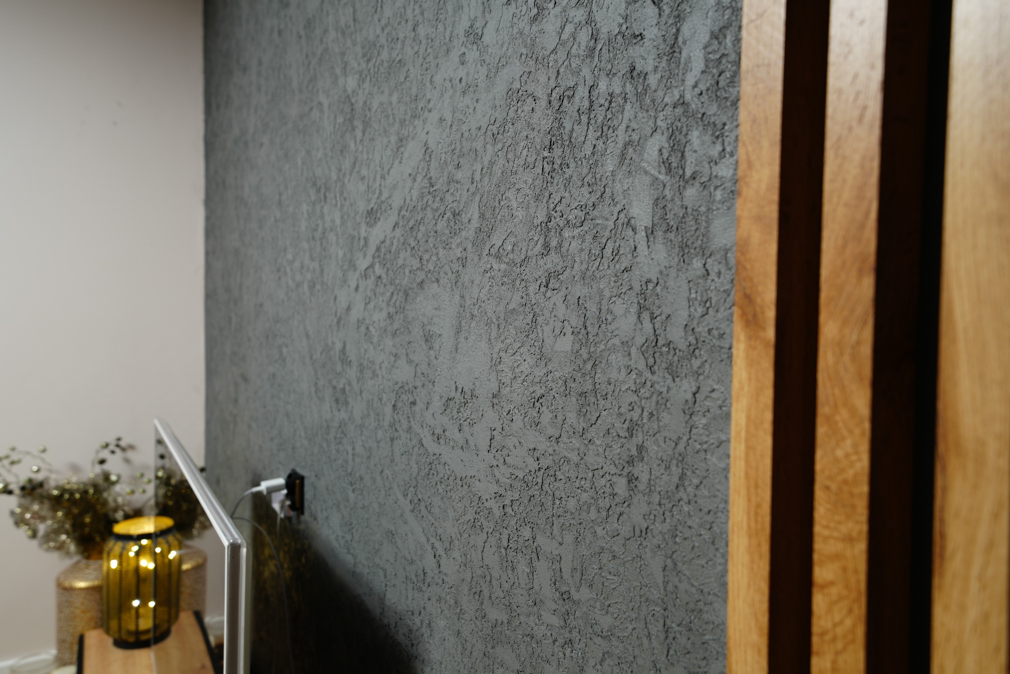 Dekoracyjna, szara ściana z efektem strukturalnym we Wrocławiu, sąsiadująca z drewnianymi panelami. Widoczny fragment telewizora i dekoracji.