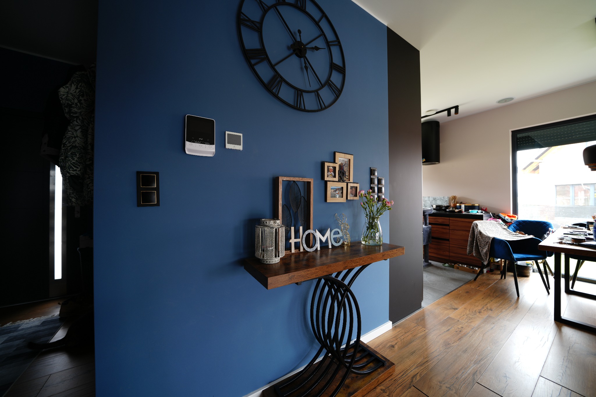 Niebieska ściana z dekoracjami: zegar, zdjęcia, konsola z napisem HOME. W tle widoczna jadalnia i parkiet na podłodze. Aranżacja wnętrza w nowoczesnym stylu.