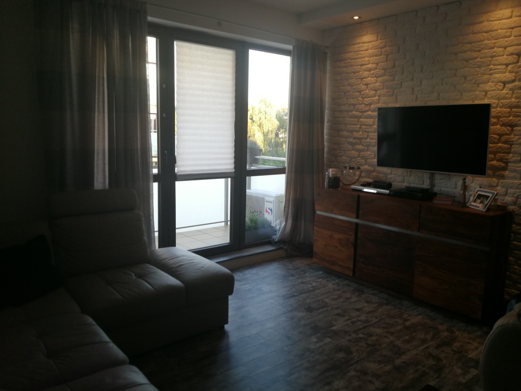 Stylowe wnętrze salonu z ceglaną ścianą, drewnianą komodą z telewizorem i wygodną sofą. Widok na balkon przez duże okno z roletami.