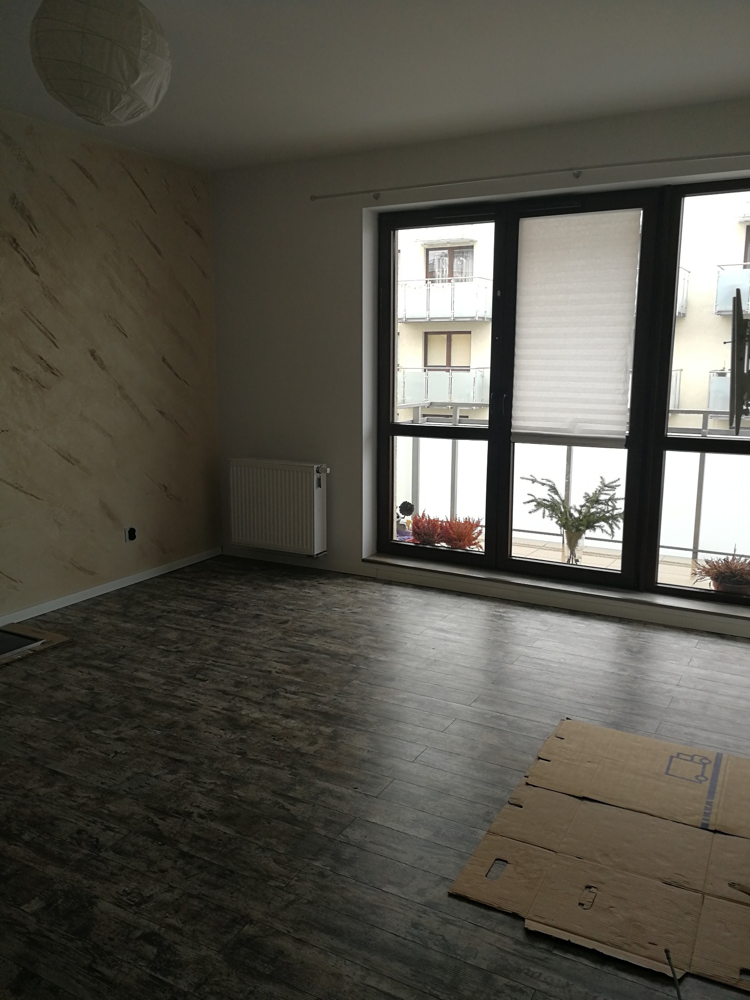 Nowoczesne wnętrze z panelami podłogowymi w odcieniach szarości i beżową ścianą z dekoracyjnym wzorem. Okno z widokiem na budynek.