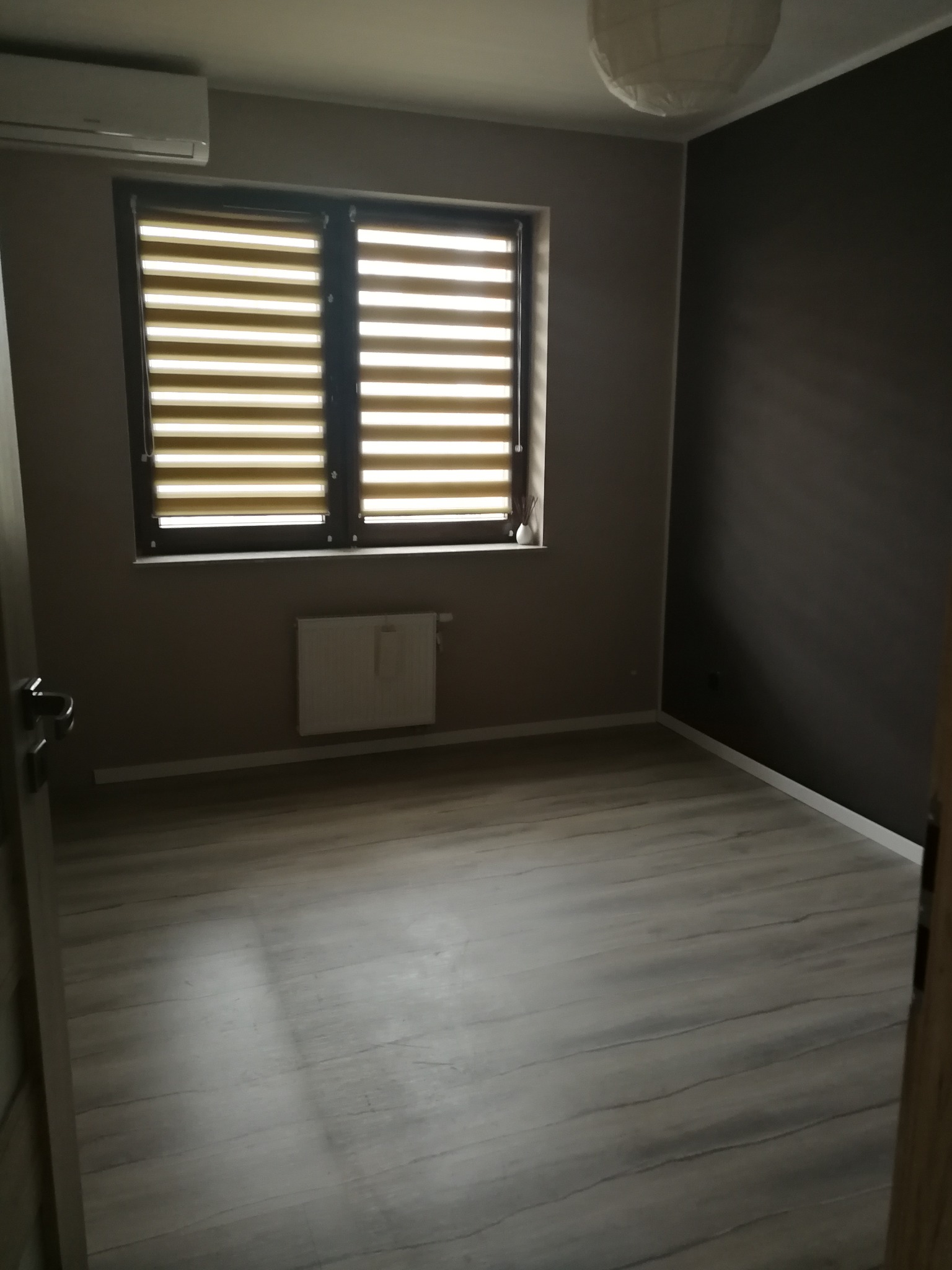 Pokój po odświeżeniu: szare ściany, drewnopodobna podłoga, żaluzje w oknie. Widoczny grzejnik i lampa sufitowa. Ciemniejsza ściana w rogu dodaje kontrastu.