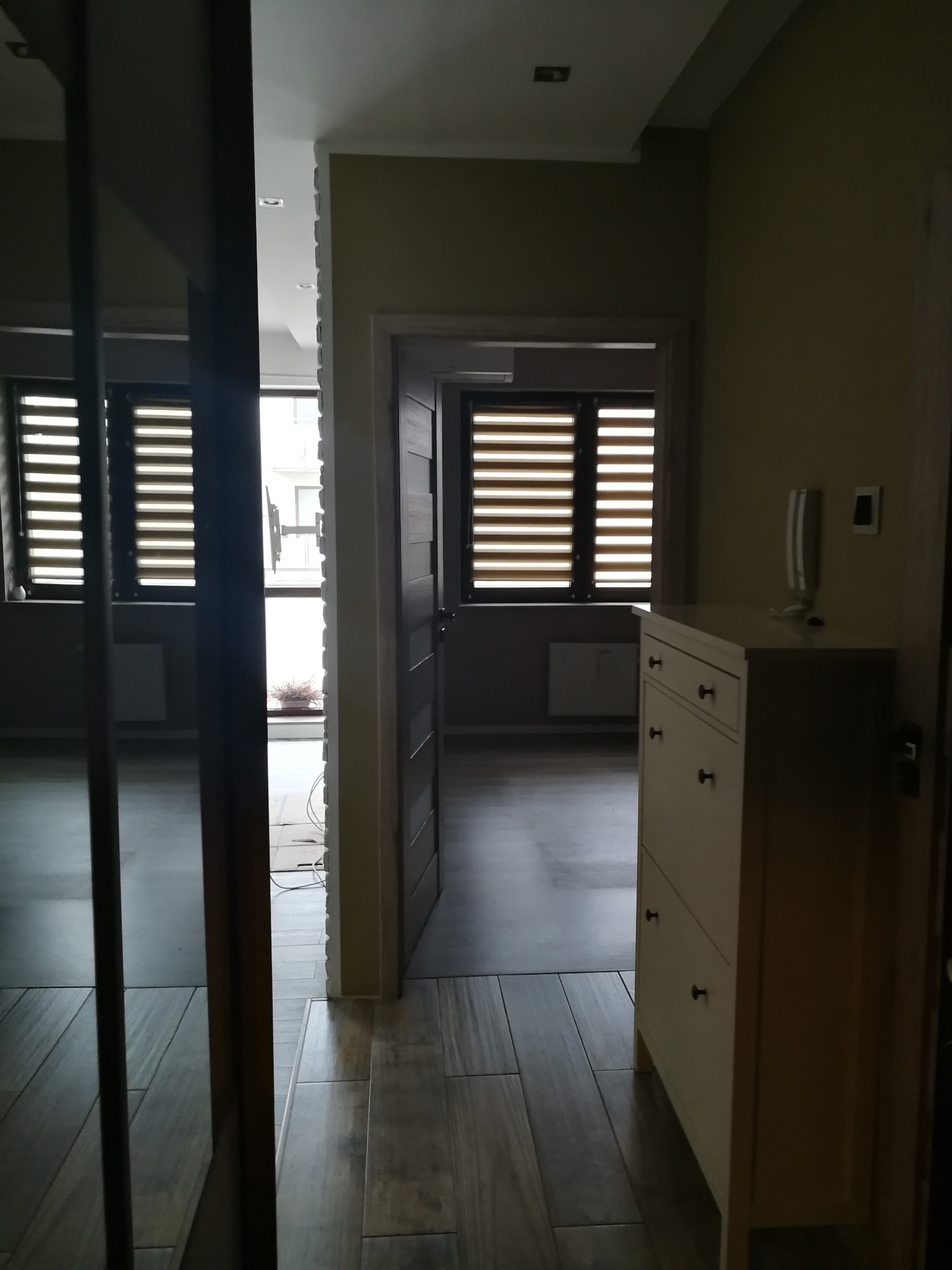 Ukończone wnętrze: przedpokój z szafką na buty w jasnym odcieniu, widok na pokój z drewnianą podłogą i oknem z żaluzjami. Minimalistyczny design.