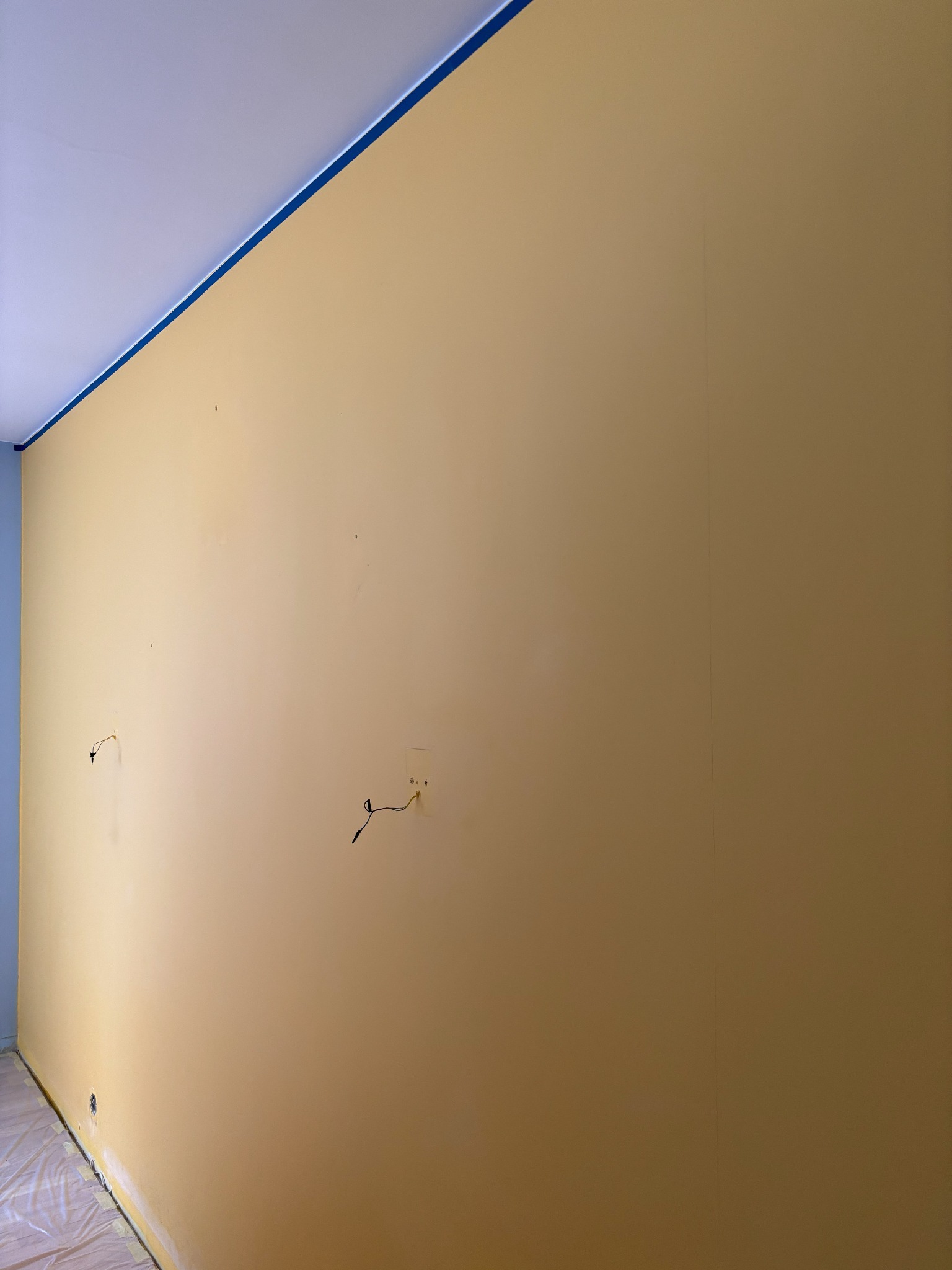 Żółta ściana po malowaniu, zabezpieczona taśmą malarską przy suficie i folią na podłodze. Widoczne gniazdka elektryczne i kable. Świeżo pomalowane wnętrze.