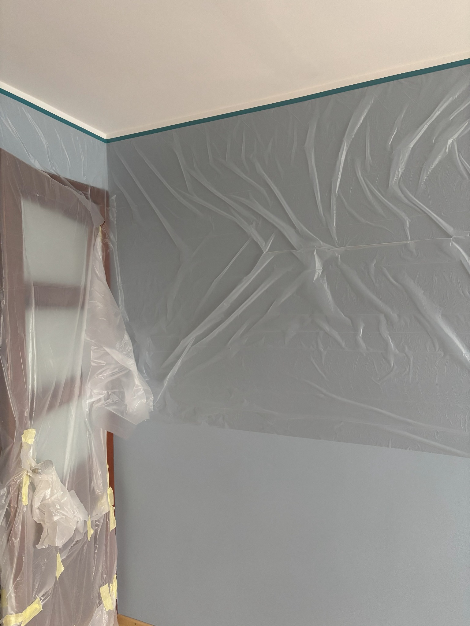 Przygotowanie do malowania: ściana i drzwi zabezpieczone folią malarską i taśmą. Ściana pomalowana na szaro, widoczny fragment sufitu z zielonym paskiem.