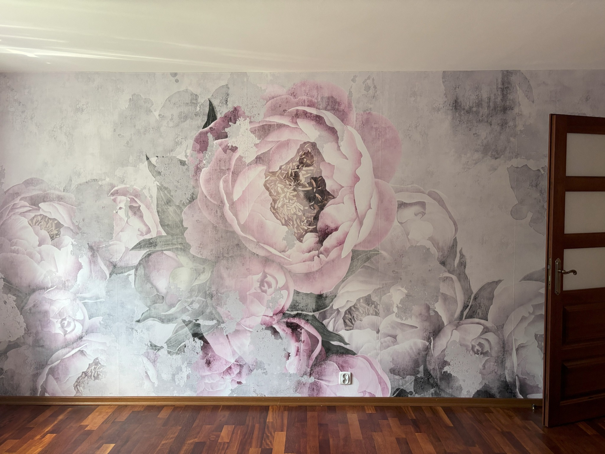 Ściana z fototapetą w kwiaty piwonii w odcieniach różu i szarości, widoczny fragment drewnianej podłogi i otwarte, drewniane drzwi.