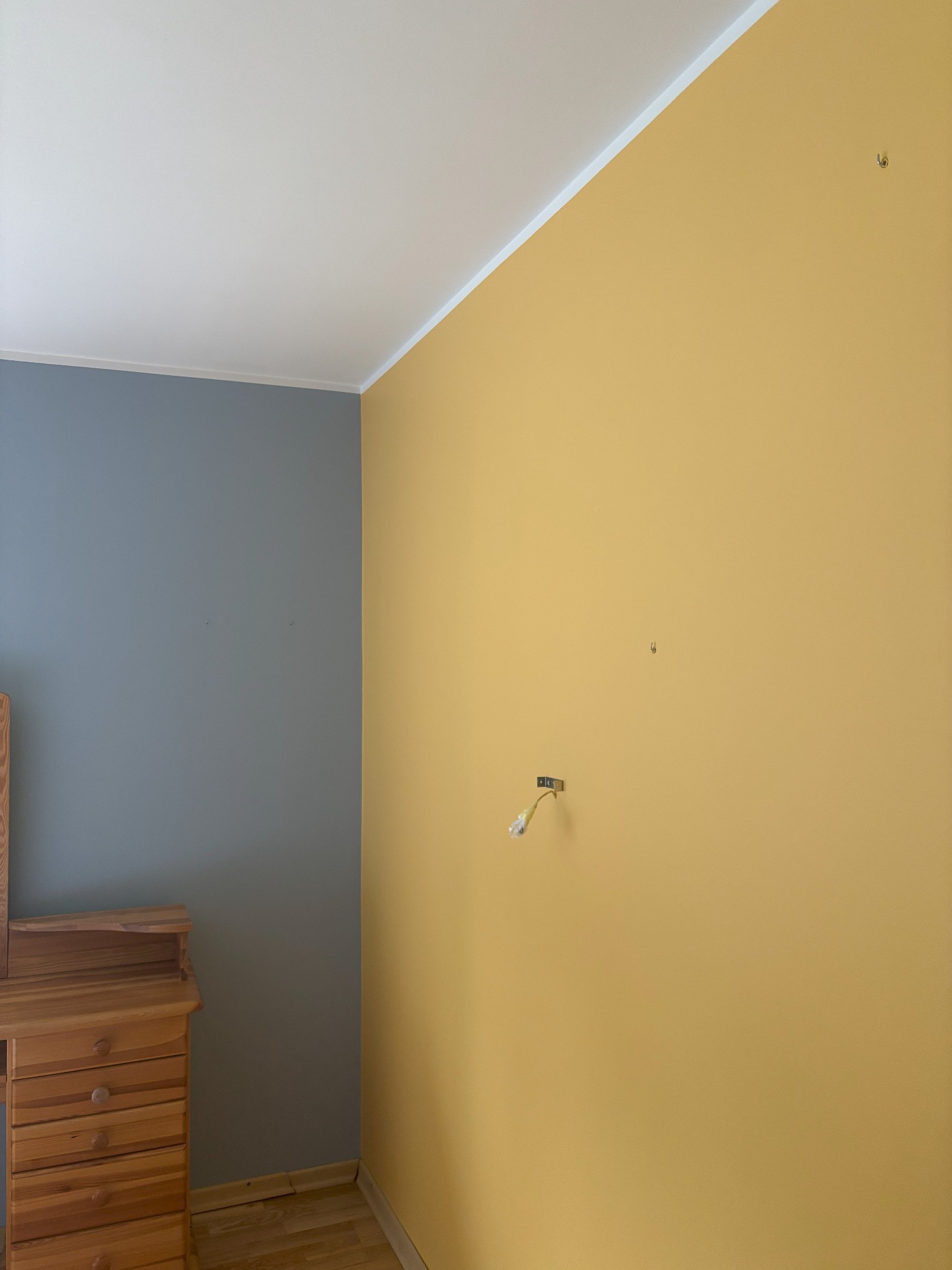 Pokój po malowaniu: ściana żółta, druga szara. Widoczny fragment drewnianej komody i wystające kable po lampie. Podłoga drewniana, jasna.