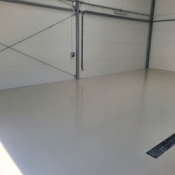 Epoxy.bp posadzki żywiczne - Jasnoszara, gładka powierzchnia w hali w Janowie Podlaskim, z kratką ściekową i wiadrem w rogu. Widoczne instalacje pod sufitem i metalowe elementy konstrukcyjne.
