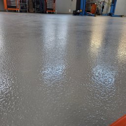 Epoxy.bp posadzki żywiczne - Szara, błyszcząca posadzka żywiczna w warsztacie samochodowym. W tle narzędzia, szafki i podnośnik. Widoczna struktura posadzki, odbijająca światło.
