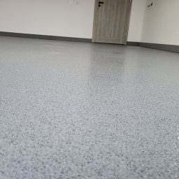 Epoxy.bp posadzki żywiczne - Jasnoszara, błyszcząca podłoga z drobnymi plamkami, zbliżenie na gładką powierzchnię w nowym wnętrzu. Widoczna listwa przypodłogowa i drzwi w tle.