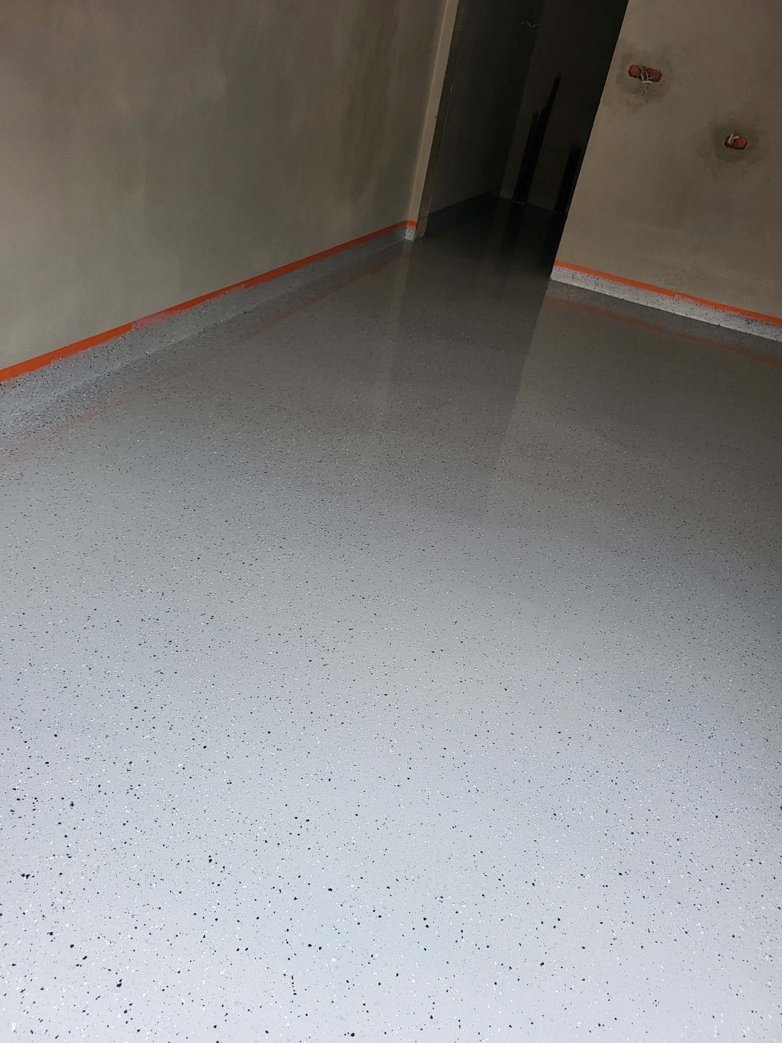Jasnoszara, błyszcząca podłoga z drobnymi, ciemnymi plamkami, zabezpieczona pomarańczową taśmą przy ścianach. Widok na korytarz w głębi. Świeżo wylana posadzka.