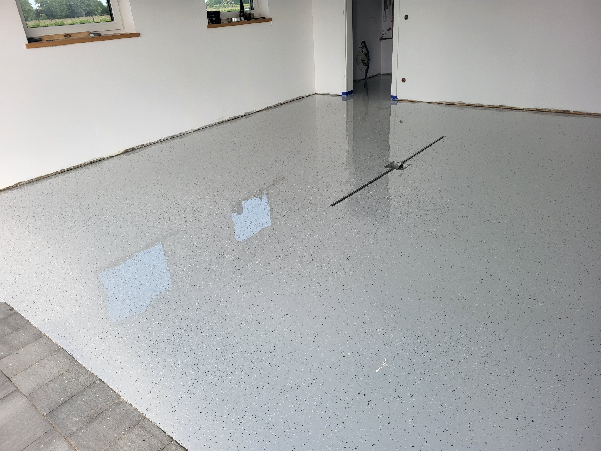 Jasnoszara, błyszcząca posadzka z odpływem liniowym w nowym wnętrzu. Widoczne okna i ślady taśmy malarskiej przy ścianach. Minimalistyczny design, gładka powierzchnia.