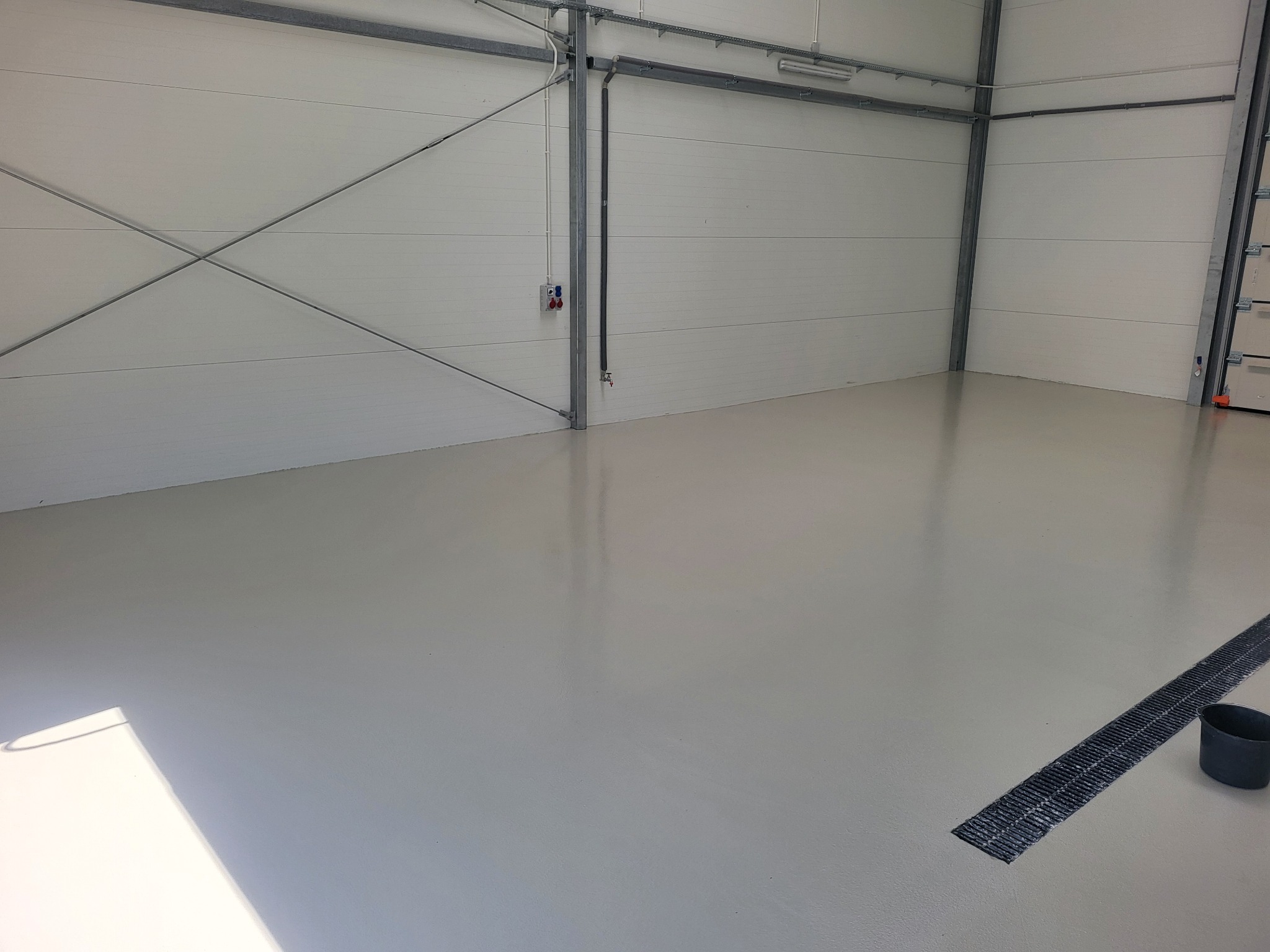 Jasnoszara, gładka powierzchnia w hali w Janowie Podlaskim, z kratką ściekową i wiadrem w rogu. Widoczne instalacje pod sufitem i metalowe elementy konstrukcyjne.