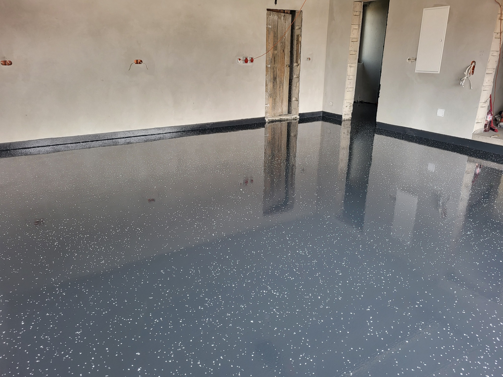 Błyszcząca, ciemnoszara posadzka z białymi drobinkami w surowym wnętrzu, z widocznymi instalacjami elektrycznymi i otworami drzwiowymi bez ościeżnic.