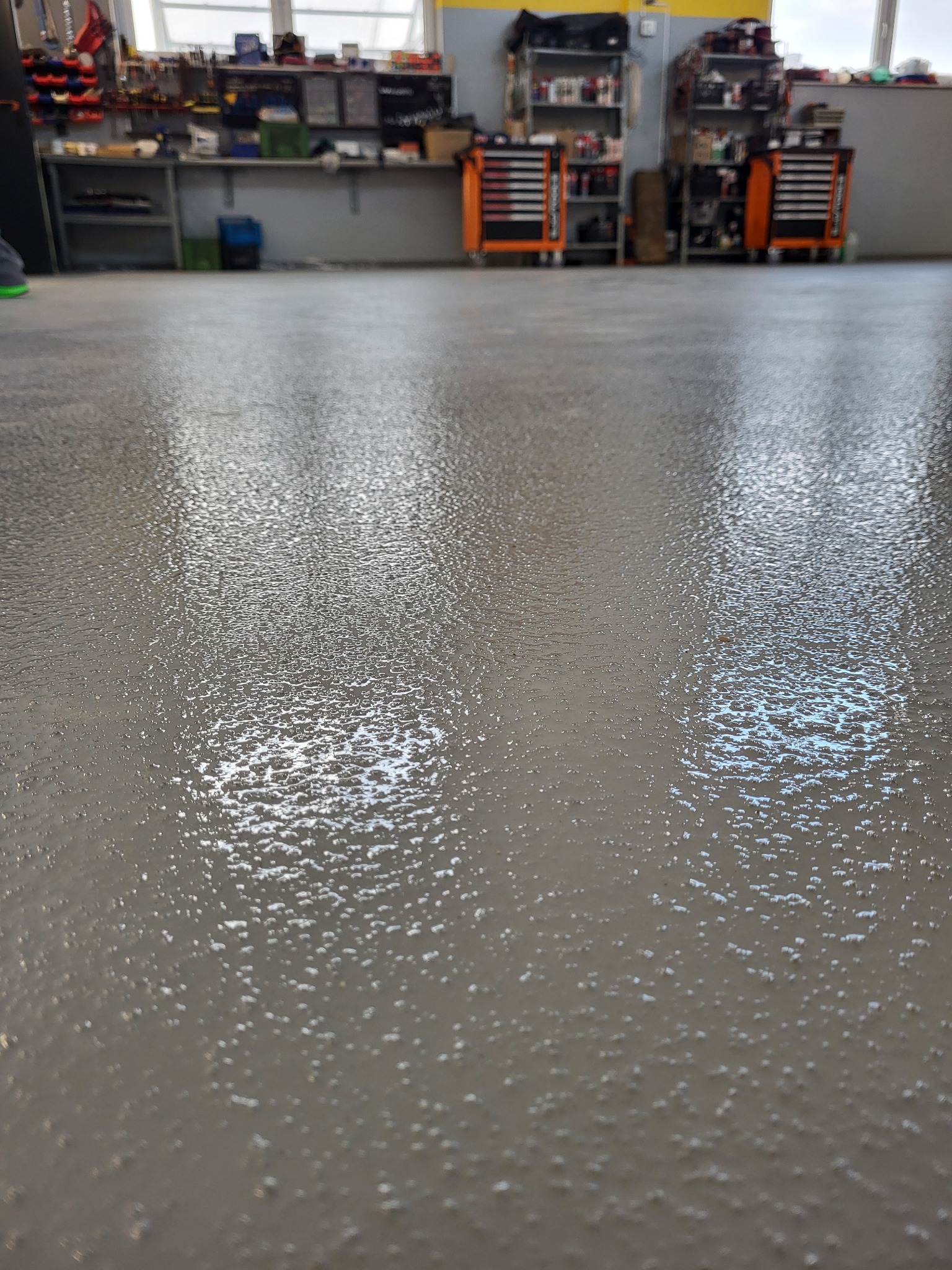 Szara, błyszcząca posadzka w warsztacie z widocznymi narzędziami i szafkami w tle. Delikatna faktura powierzchni. Widok z niskiej perspektywy.