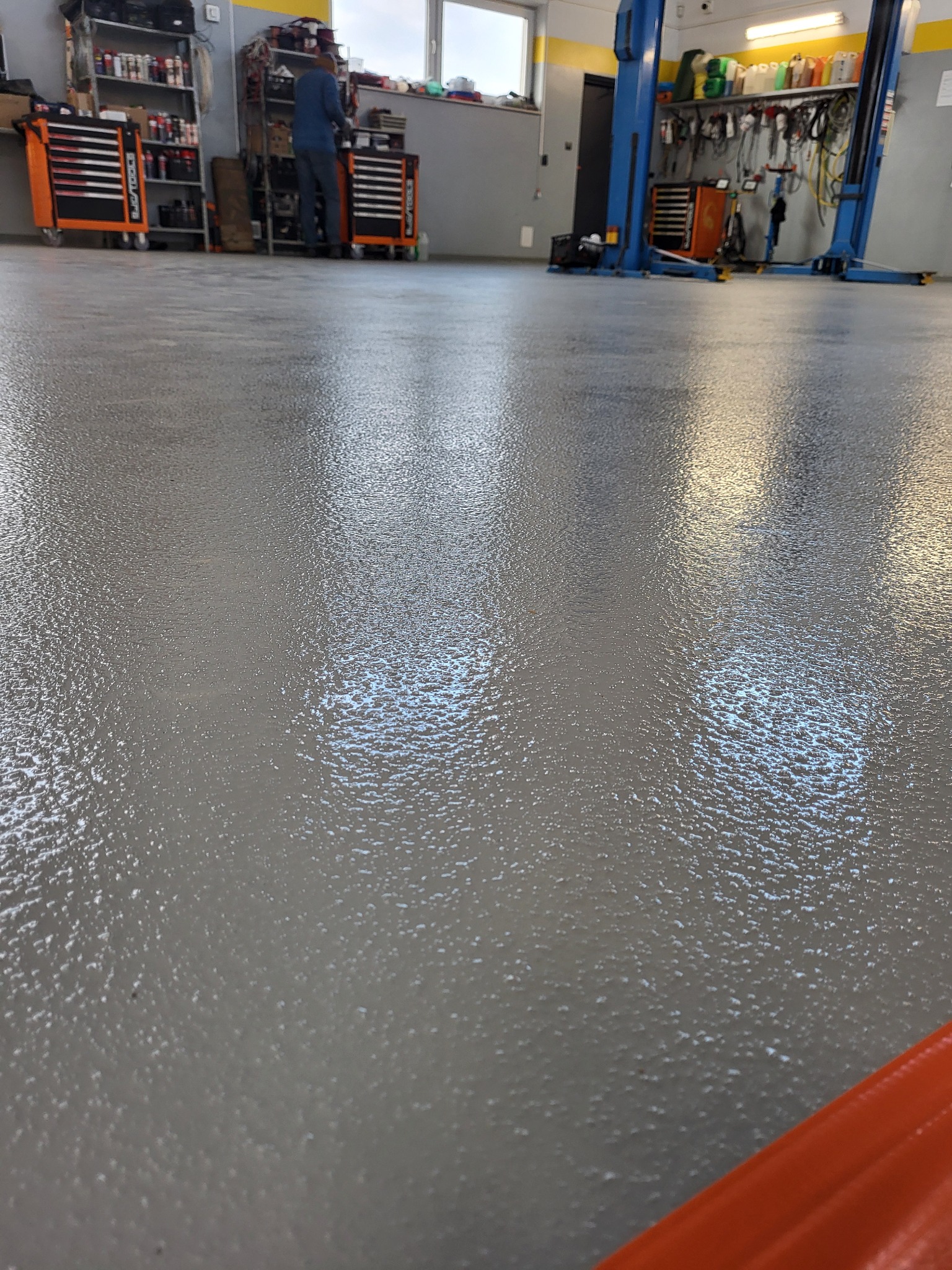 Szara, błyszcząca posadzka żywiczna w warsztacie samochodowym. W tle narzędzia, szafki i podnośnik. Widoczna struktura posadzki, odbijająca światło.