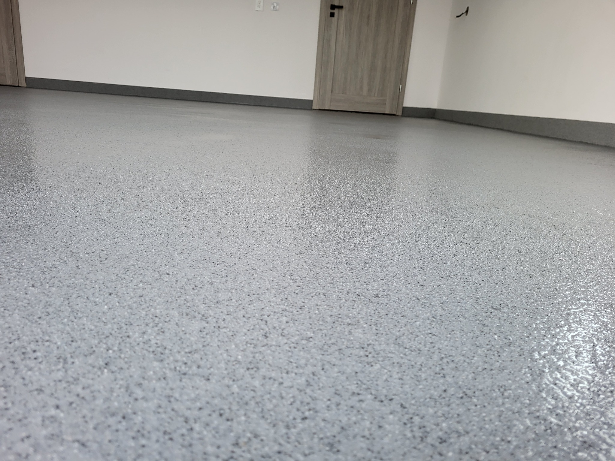 Jasnoszara, błyszcząca podłoga z drobnymi plamkami, zbliżenie na gładką powierzchnię w nowym wnętrzu. Widoczna listwa przypodłogowa i drzwi w tle.