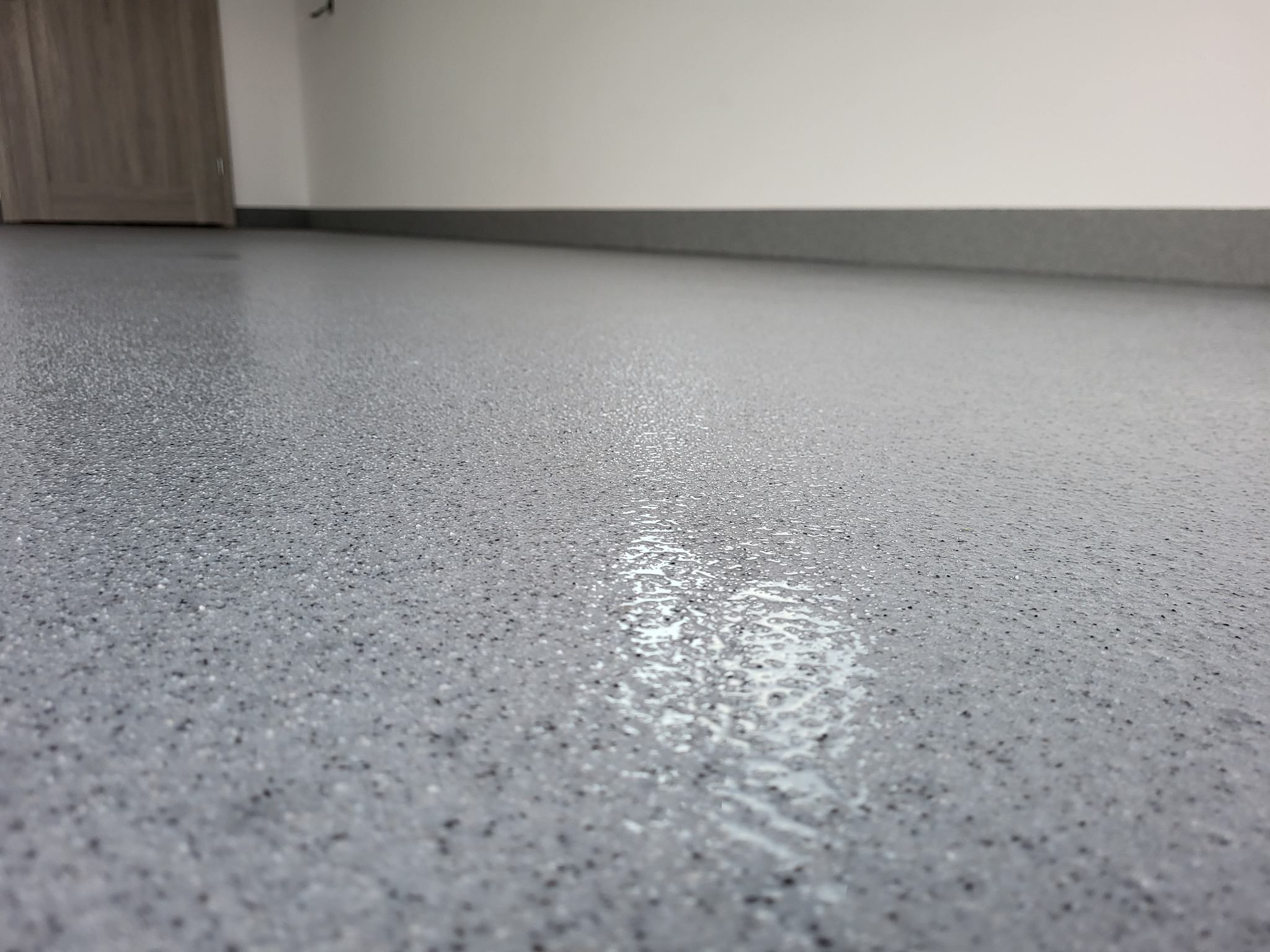Gładka, szara posadzka z drobnymi czarnymi plamkami, odbijająca światło. W tle widoczna ściana i fragment drzwi.