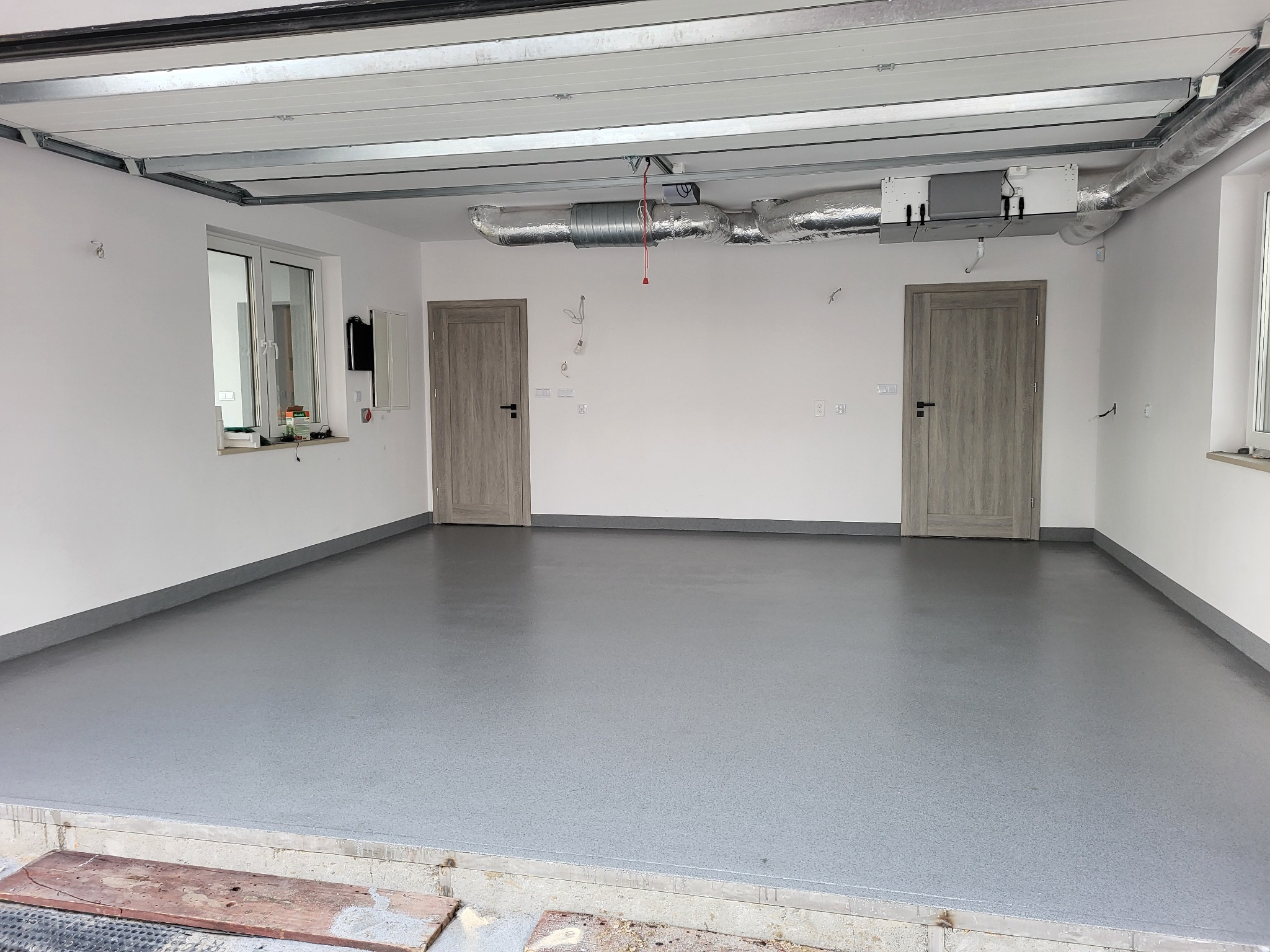 Wykończone wnętrze garażu z szarą, jednolitą podłogą. Widoczne dwie drewniane drzwi, białe ściany, okno i instalacja wentylacyjna pod sufitem.