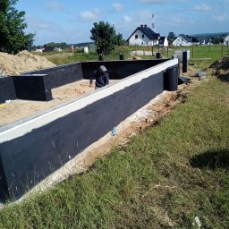MUR-BET Kazimierz Reszka - Fundamenty domu w trakcie hydroizolacji, widoczny pracownik z narzędziem, teren budowy, w tle zabudowa jednorodzinna i błękitne niebo.
