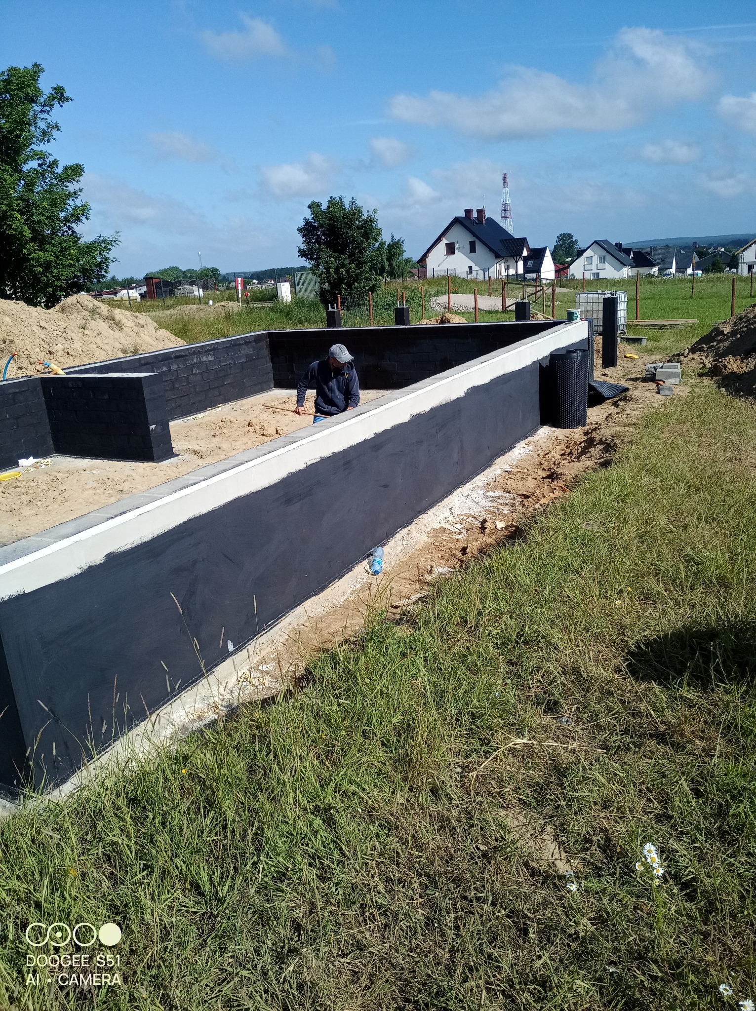 Fundamenty domu w trakcie hydroizolacji, widoczny pracownik z narzędziem, teren budowy, w tle zabudowa jednorodzinna i błękitne niebo.