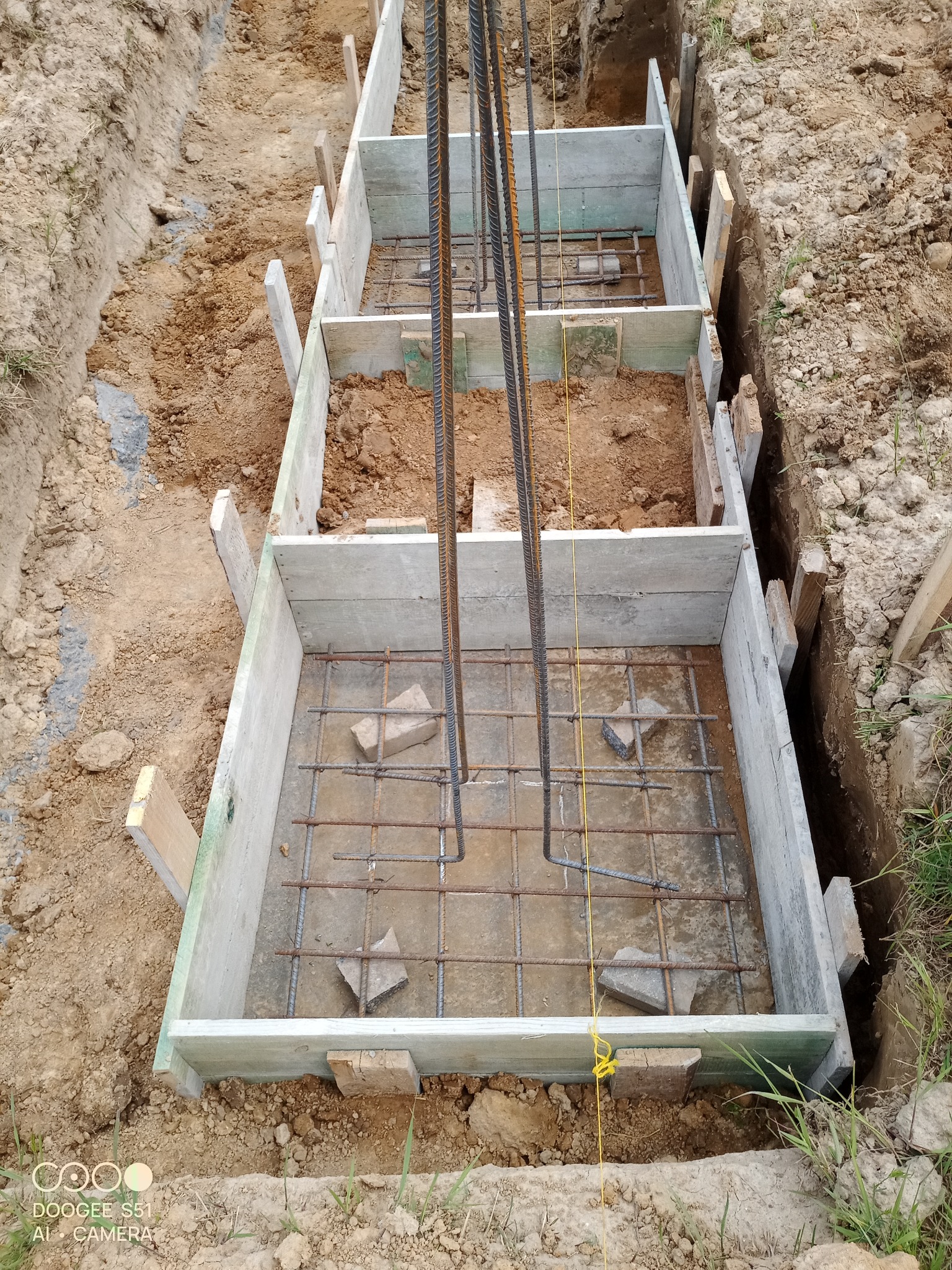 Szczegółowe ujęcie z góry: szalunek fundamentowy z siatką zbrojeniową i pionowymi prętami, przygotowany do zalania betonem w wykopie ziemnym.