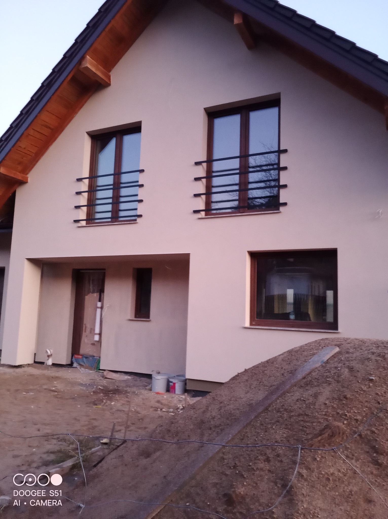 Elewacja domu jednorodzinnego w Krokowej po remoncie. Jasny budynek z brązowymi oknami i balustradami. Widoczny fragment działki z ziemią i wiadrami.