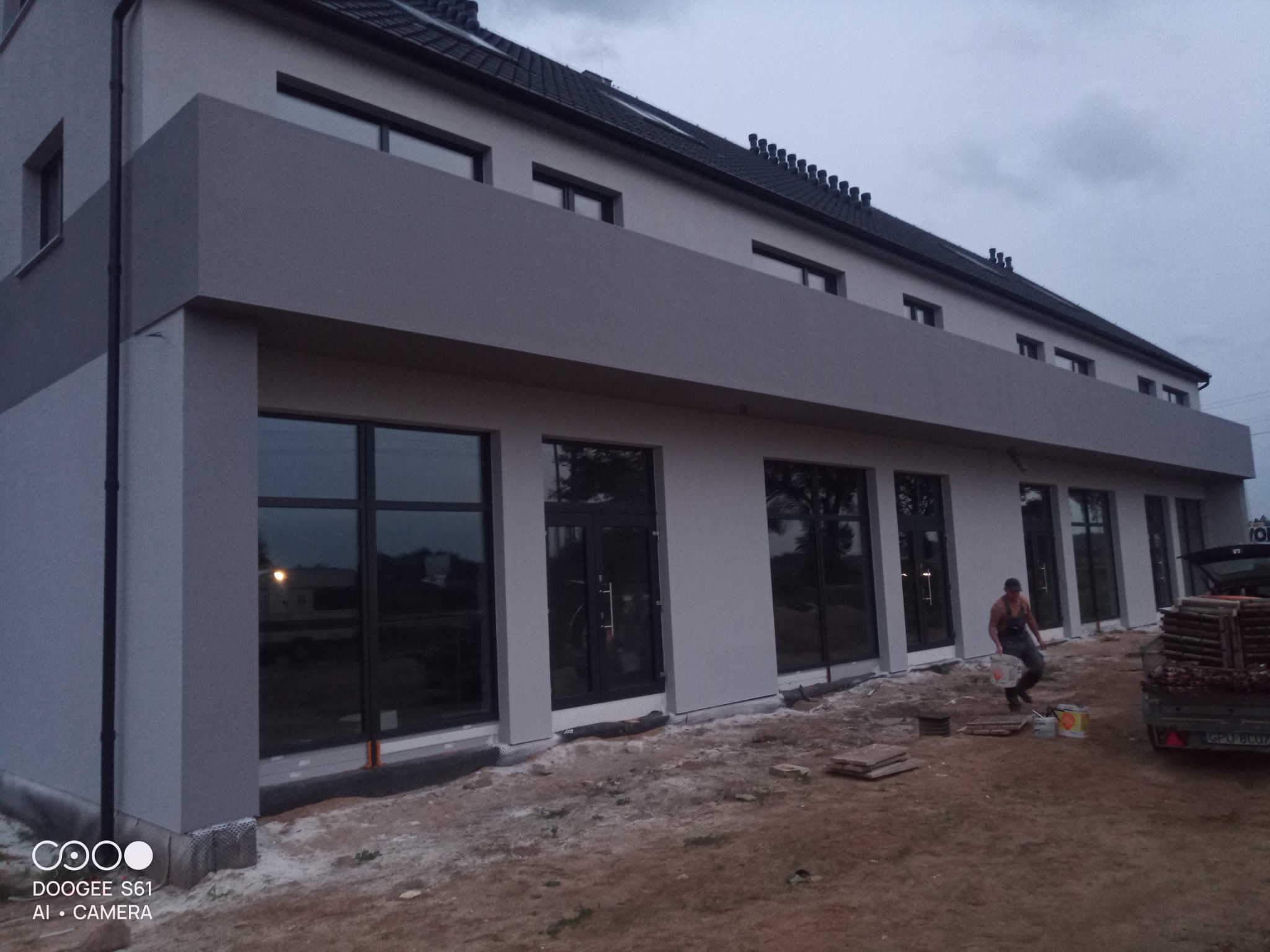 Szary budynek z rzędem okien i pracownikiem budowlanym niosącym wiadro, z widoczną przyczepą i materiałami budowlanymi. Ujęcie z dołu, pochmurne niebo.