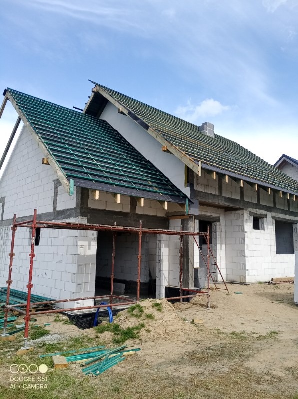 Dom w trakcie budowy: białe ściany z bloczków, zielona konstrukcja dachu, rusztowania. Widok z zewnątrz, w tle błękitne niebo.