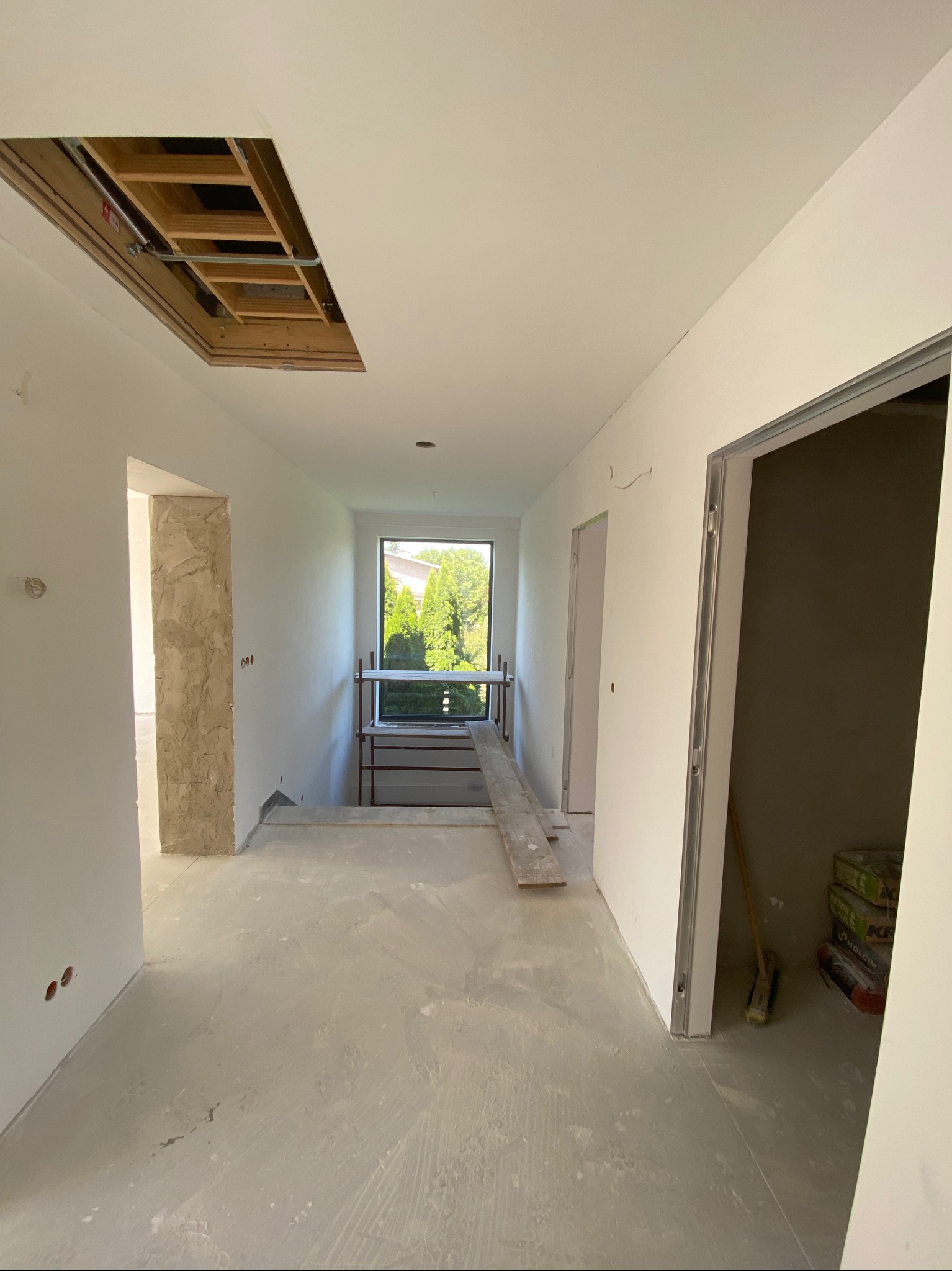 Wnętrze w trakcie prac wykończeniowych: gładkie, białe ściany, surowa podłoga, widoczne worki z zaprawą i rusztowanie przy oknie. Otwór w suficie odsłania konstrukcję.