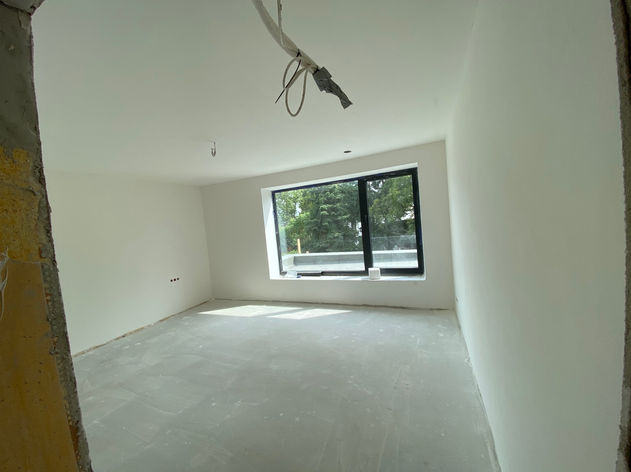Wnętrze pokoju po szpachlowaniu i malowaniu na biało, widoczne gładkie ściany, okno z widokiem na zieleń i przygotowana podłoga. Fragment surowej ściany po lewej.
