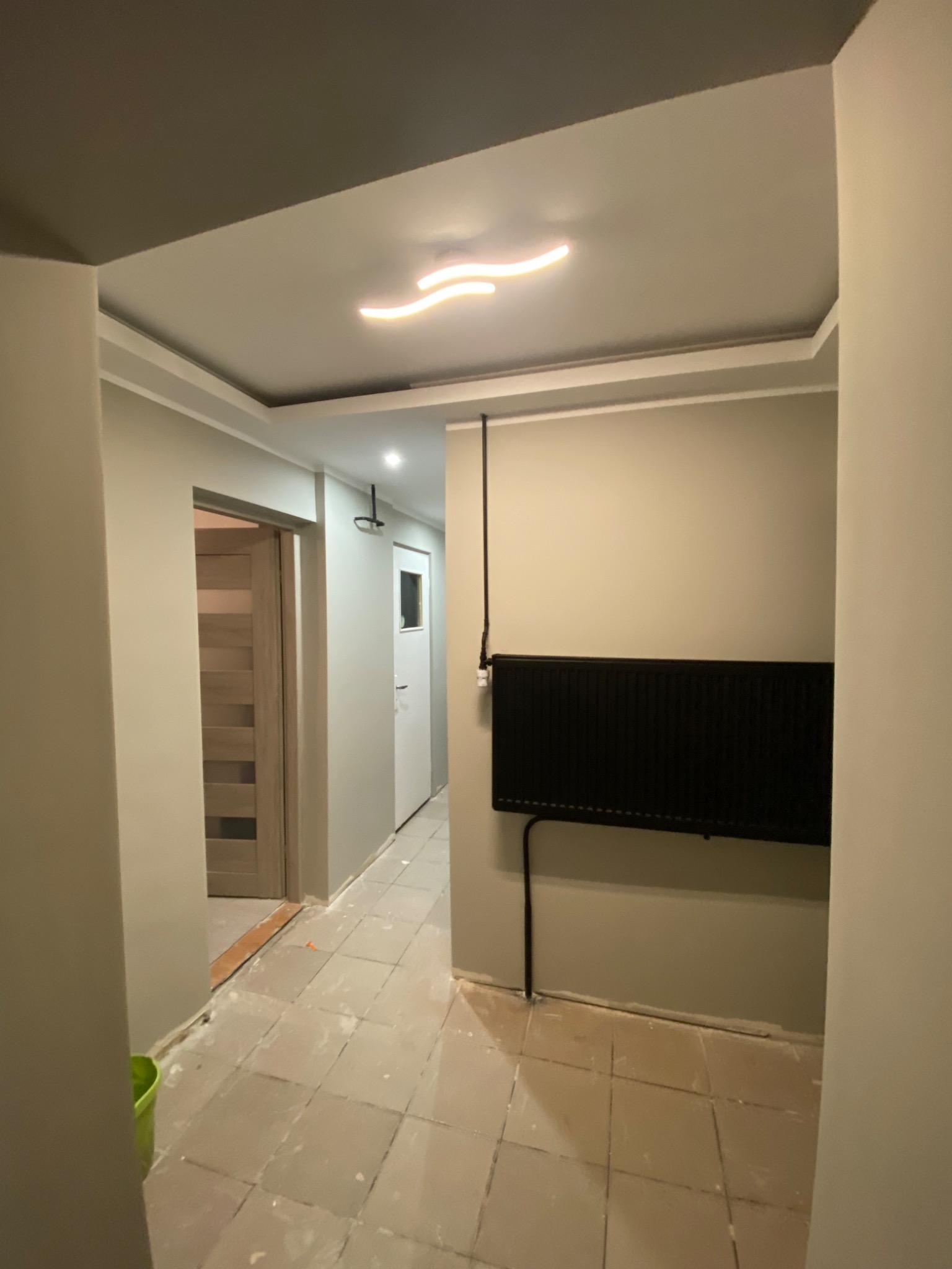 Wnętrze korytarza w trakcie remontu: szare ściany, czarny grzejnik, widoczne ślady prac na podłodze. Nowoczesne oświetlenie sufitowe w kształcie fali. Drzwi widoczne w tle.