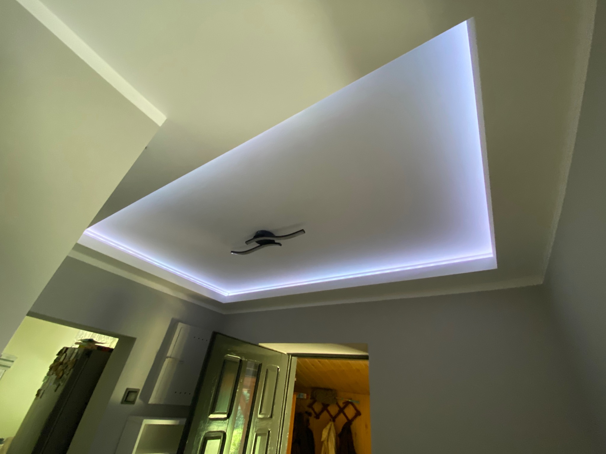 Podwieszany sufit z oświetleniem LED w kształcie trapezu, widok z dołu, nowoczesny design, czarna lampa sufitowa na środku, ściany w odcieniach szarości.