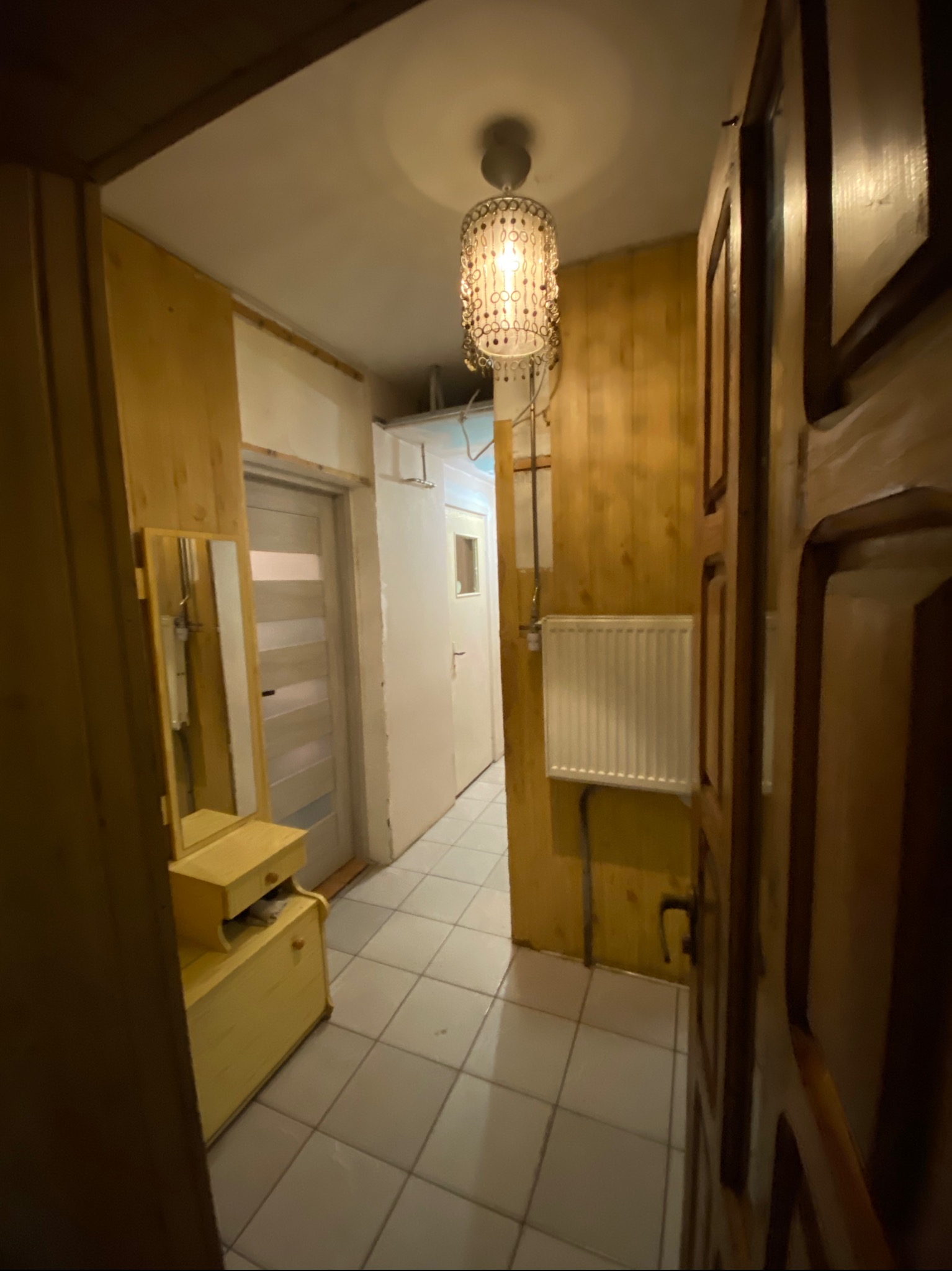 Korytarz z boazerią i grzejnikiem, oświetlony ozdobną lampą. Widoczna szafka z lustrem i płytki na podłodze. Ujęcie z perspektywy wejścia do pomieszczenia.