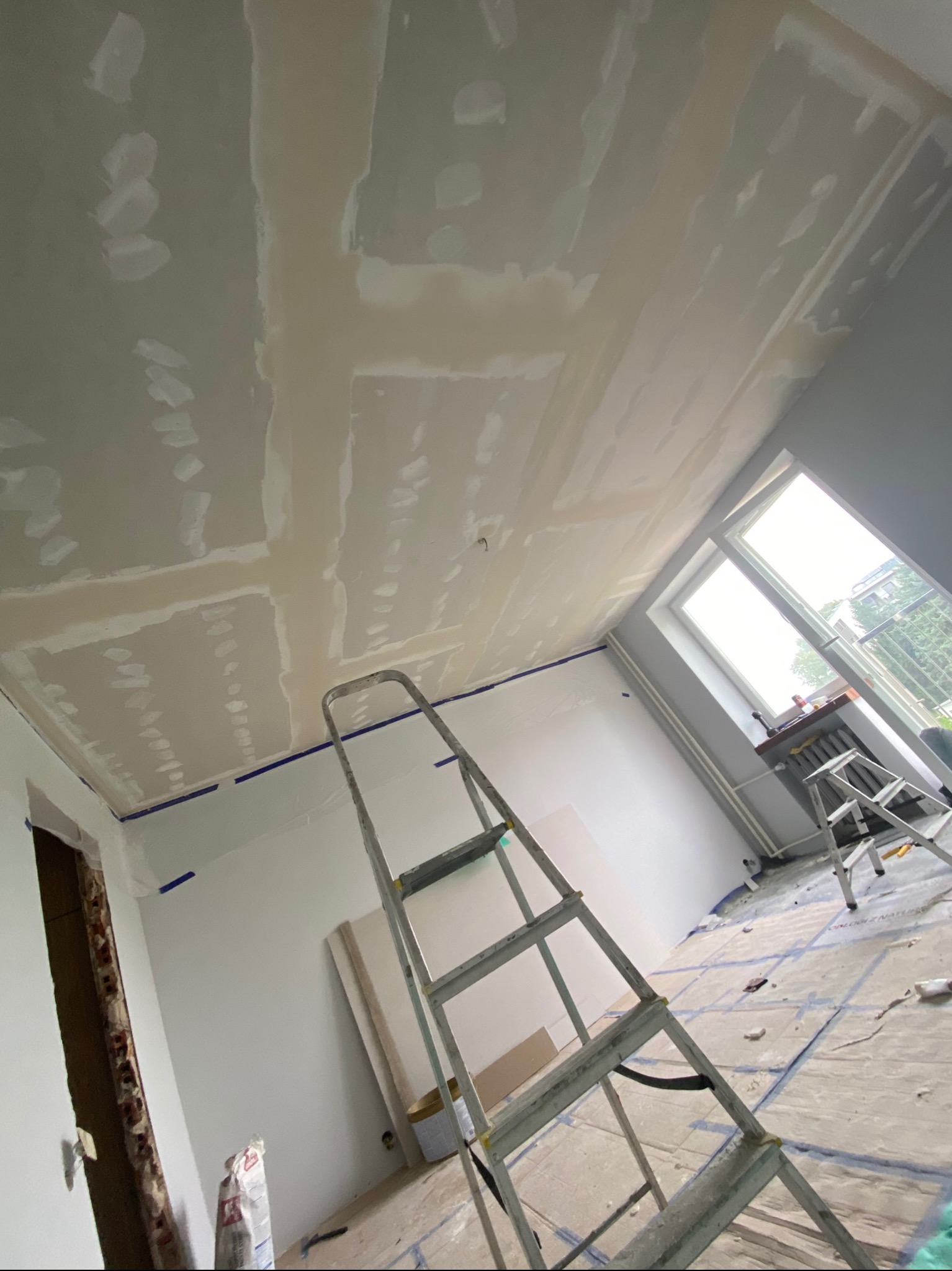 Pokój w trakcie remontu: sufit z widocznymi łączeniami płyt kartonowo-gipsowych, drabina, zabezpieczona podłoga. Etap szpachlowania przed malowaniem.