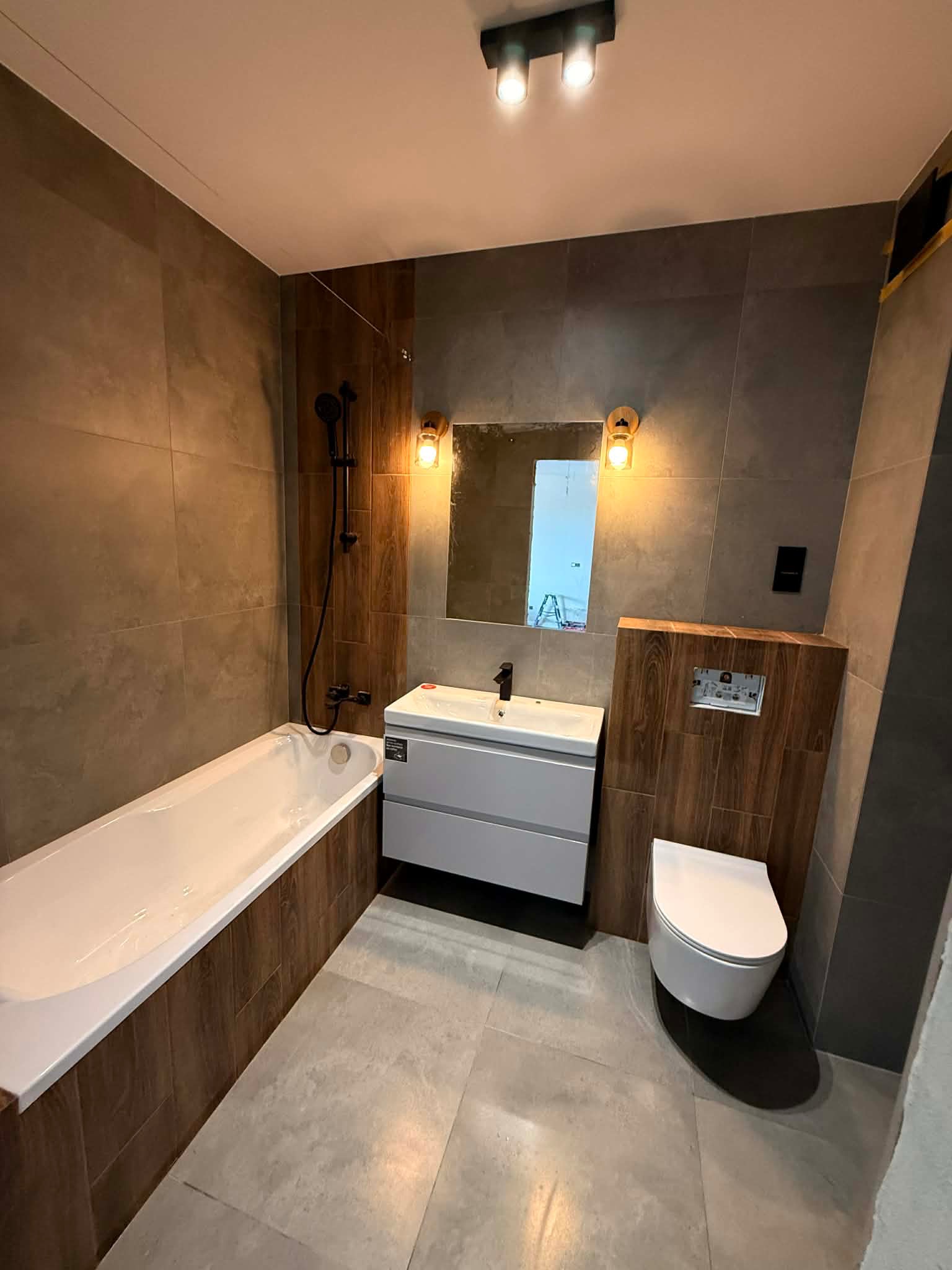 Nowoczesna łazienka po remoncie: beżowe płytki, wanna z drewnopodobną obudową, biała umywalka z szafką i toaleta podwieszana. Oświetlenie punktowe.