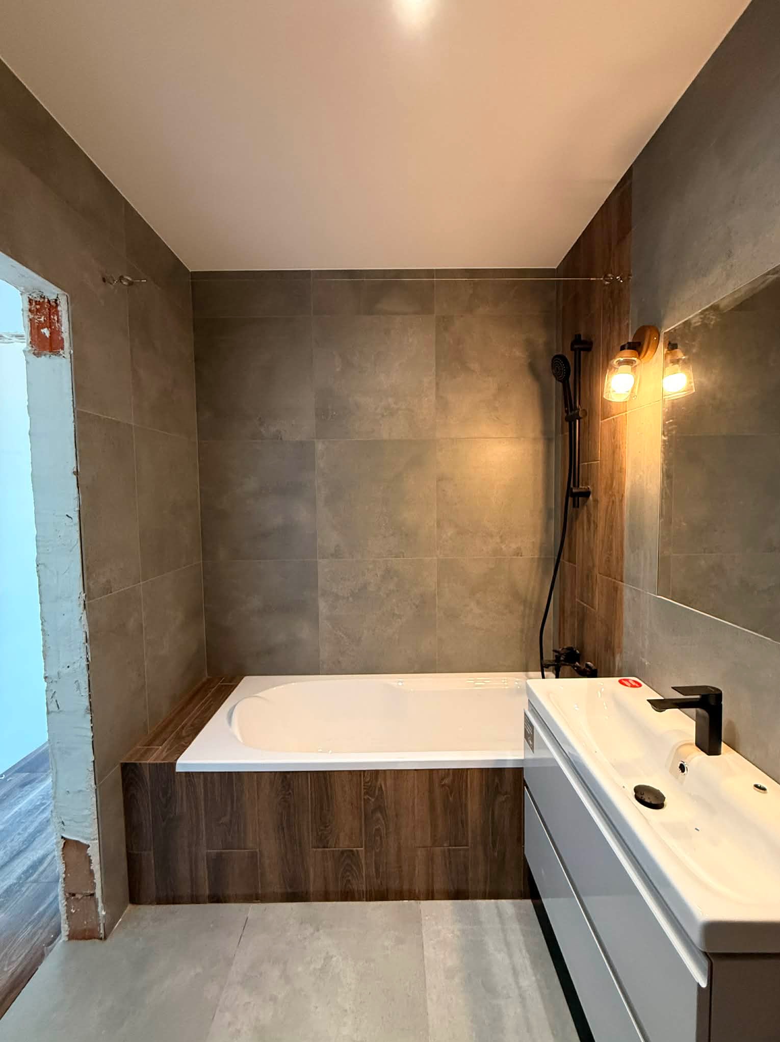 Wykończona łazienka z wanną obudowaną drewnopodobnymi panelami, szarą glazurą i białą szafką z umywalką. Widoczna czarna armatura i oświetlenie w stylu retro.