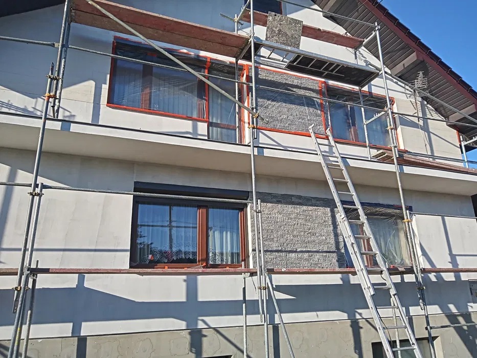 Dom w trakcie prac elewacyjnych: fragment ściany z nałożonym kamieniem dekoracyjnym, rusztowanie i drabina. Okna zabezpieczone folią.