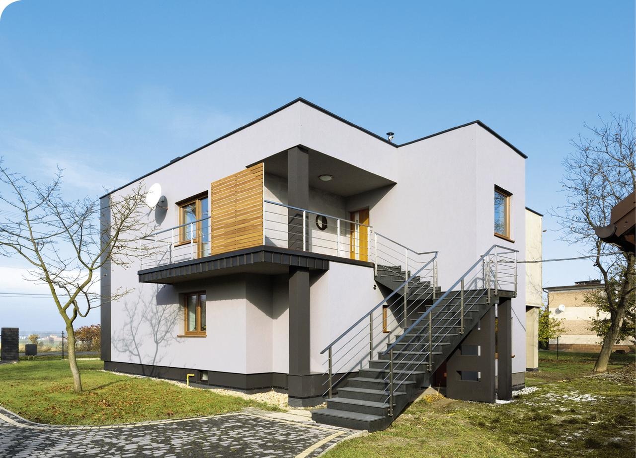 Nowoczesny dom z szarą elewacją, balkonem z drewnianą przesłoną i zewnętrznymi schodami. W tle błękitne niebo i zieleń trawnika.